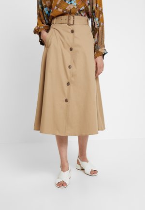 Vrouw draagt een beige, knoopsluitende A-lijn midi-rok met een brede riem, gecombineerd met een lange mouwen blouse met patroon en witte sandalen met hak.