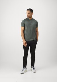 Férfi rövid ujjú polo ing sötétzöld színben, fél zippes gallérral és logós részletekkel, párosítva fekete slim fit nadrággal és fehér tornacipővel.