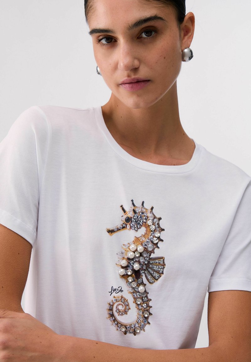 Γυναίκα που φοράει λευκό t-shirt με λεπτομερή σχεδιασμό ιππόκαμπου με πετράδια και μεγάλα ασημένια σφαιρικά σκουλαρίκια σε απλό φόντο.