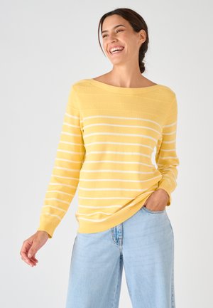 Vrouw die glimlacht, draagt een gele trui met lange mouwen en witte strepen en lichtblauwe jeans, met één hand in haar zak, tegen een effen achtergrond.