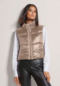 Beige, gesteppte Pufferweste mit Stehkragen und durchgehendem Reißverschluss. Getragen über einem weißen Langarmshirt, kombiniert mit schwarzen Hosen. Glattes, glänzendes Material.