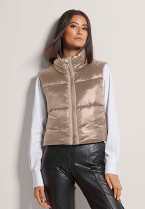 Beige quilted puffer vest with a mock neck and front zipper. Worn over a white long-sleeve shirt, paired with black pants. Smooth, shiny fabric.