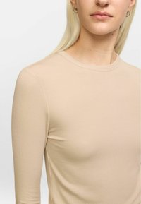 Beige ribbed long-sleeve top with a round neckline. Soft texture, form-fitting design, and subtle seams along the shoulders.