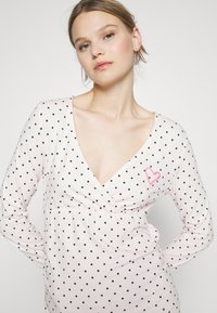 Långärmad rosa topp med svarta prickar, V-utsnitt och en rynkad midja. Har en liten rosa hjärtdekoration på bröstet.