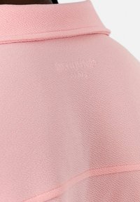 Ljust rosa strukturerad tyg med krage och präglad logodetalj. Det mjuka materialet har subtila mönster och sömnadsdetaljer.