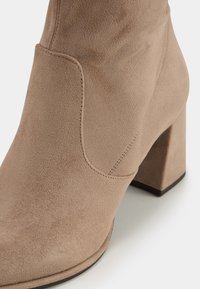 Suede ankle boot in light beige with a square-shaped heel, stitched detailing, and a side cutout for breathability and style.
