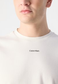 Balta kokvilnas T-kreklu ar apaļu kakla izgriezumu un melnu "Calvin Klein" logo izšuvumu krūtīs. Gluda tekstūra.