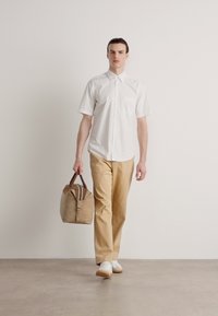Camisa blanca de manga corta con botones, pantalones beige y zapatillas blancas. Lleva una bolsa beige con detalles marrones, de pie sobre un suelo de hormigón.