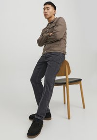 Giacca button-up marrone con tasche, jeans grigio scuro e scarpe nere, seduto su una sedia di legno con seduta nera. Sfondo minimalista.