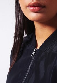 Námořnická bomber bunda s hladkým, lesklým povrchem, zapínáním na zip a žebrovaným límcem. Detail krku a rtů na neutrálním pozadí.