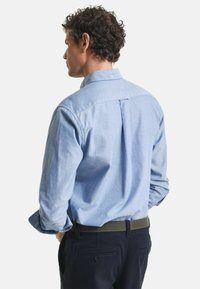 Lichtblauw overhemd met een puntkraag, lange mouwen en een rugplooitje. Draagt het met donkere broek en een groene riem met textuur.