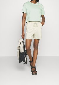 Jasnozielony krótki t-shirt, beżowe szorty na sznurku, czarne sandały oraz beżowo-czarny plecak. Prosty design, luźny krój.