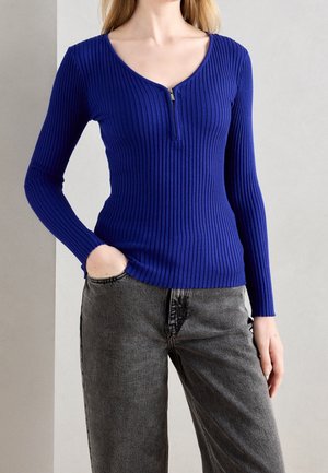 Frau trägt ein eng anliegendes, geripptes, langärmeliges blaues Oberteil mit einem Reißverschluss vorne, kombiniert mit grauen, hoch taillierten Jeans, Hand in der Tasche.
