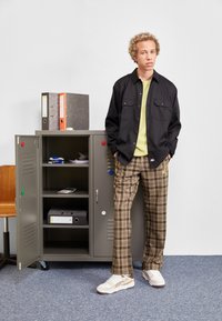 Chemise noire boutonnée sur une chemise vert clair, pantalon cargo à carreaux marron et baskets blanches. Casier gris et fournitures de bureau en arrière-plan.