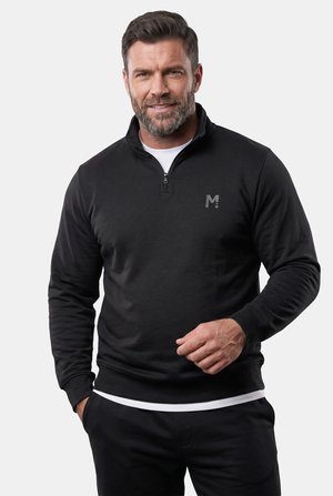 Mann mit kurzem Haar und Bart trägt ein schwarzes Quarter-Zip-Sweatshirt über einem weißen Shirt, steht und lächelt vor einem schlichten hellen Hintergrund.