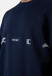 Sweatshirt bleu marine avec col rond, arborant des logos brodés blancs : initiales stylisées, "ESTEEM", et design ornemental sur la manche gauche.