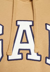 Sweat à capuche beige avec une capuche spacieuse, arborant un grand logo « GAP » brodé en blanc et bleu marine. La texture est lisse avec des coutures visibles.