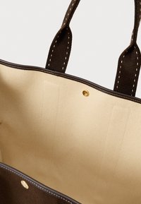 Εσωτερικό από μπεζ ύφασμα τσάντας tote με σκούρο καφέ ραμμένα χερούλια και άκρες, διαθέτοντας ένα μικρό χρυσό κούμπωμα με κουμπί.