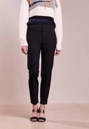 Person trägt schwarze, maßgeschneiderte Knöchelhosen, gestreiften Strickpullover mit Fransen und schwarze offene High Heels mit metallischen Knöchelriemen.