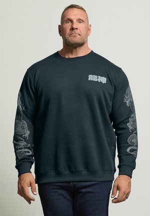 Homme portant un sweat-shirt noir avec des motifs de dragons blancs sur les deux manches et des caractères asiatiques blancs sur la poitrine gauche, debout devant un fond uni.