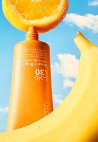Tubo de crema solar naranja posicionado en vertical, con un color naranja brillante, etiqueta SPF 30, con un plátano y una naranja encima.