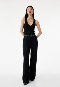 Zwarte geribbelde tanktop met een diepe V-hals en drie knopen, gecombineerd met hoog-waisted zwarte wijde broek en een zwarte riem.