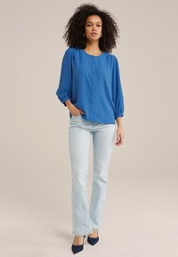 Blusa blu con bottoni, dotata di un pattern a pois testurizzati, maniche arricciate e scollatura rotonda, abbinata a jeans azzurri svasati e tacchi blu navy.