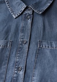 Camicia in denim con colletto a punta, bottoni frontali e due tasche sul petto. Il tessuto è in un lavaggio scuro con una sottile texture e cuciture a contrasto.
