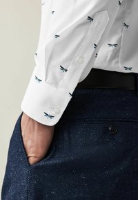 Persona vestida con una camisa blanca con estampados de libélulas azules y pantalones oscuros texturizados azul marino, con una mano casualmente en el bolsillo.