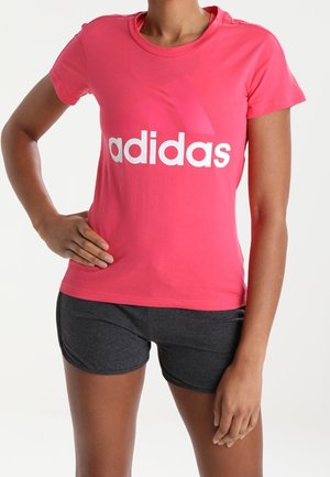 Žena v jasně růžovém tričku Adidas a tmavě šedých šortkách stojí s jednou rukou na boku před jednobarevným pozadím.