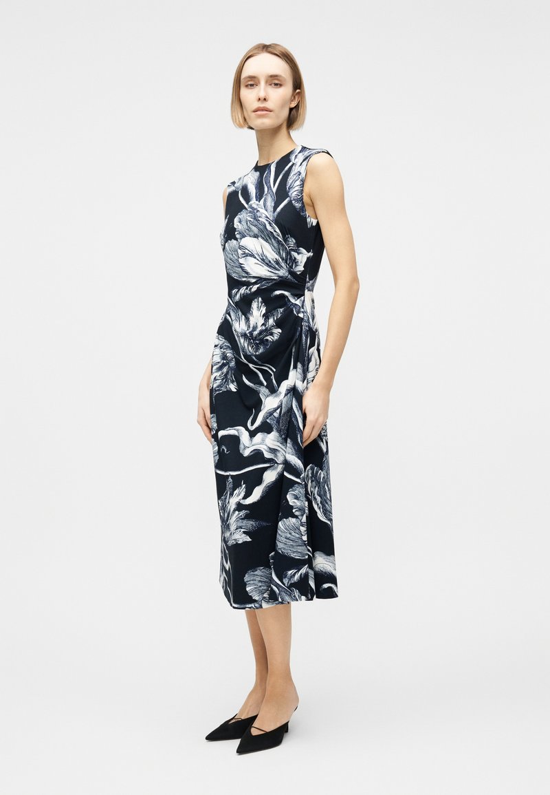 Woman wearing sleeveless black midi dress with large white floral print and black pointed low heels, standing against plain light background.