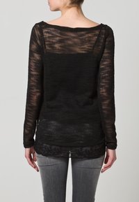 Schwarzer, langärmliger, gestrickter Pullover mit lockerem Sitz, der ein strukturiertes Design und ein geschichtetes Aussehen über einem gemusterten unteren Kleidungsstück aufweist.