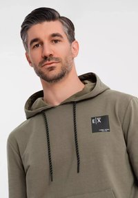Sudadera de color verde oliva con un bolsillo tipo canguro, cordones ajustables y un parche de logo negro en el pecho. Tejido suave, corte holgado.