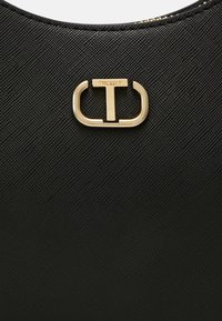 Gros plan sur un cuir noir texturé avec un logo métallique doré comportant la lettre T et le mot Twinset au-dessus.