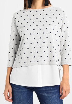 Frau trägt ein graues, langärmeliges Polka-Dot-Oberteil über einem weißen Hemd, kombiniert mit blauen Jeans und schwarzen Quasten-Ohrringen.