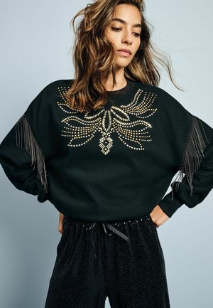 Felpa nera con decorazioni metalliche, dettagli con frange sulle maniche e vestibilità casual. Indossata con pantaloni neri scintillanti.