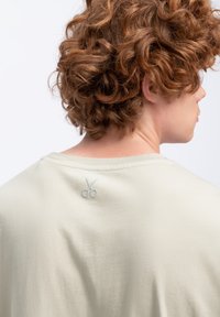 Lichtbeige T-shirt met een ronde hals, voorzien van een subtiele schaar borduursel op de achterkant van de schouder. Zachte, gladde katoenstof.