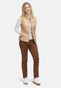 Beige gewatteerde vest met zijzakken, gedragen over een wit overhemd met lange mouwen, gecombineerd met bruine suède-achtige broek en witte sneakers.