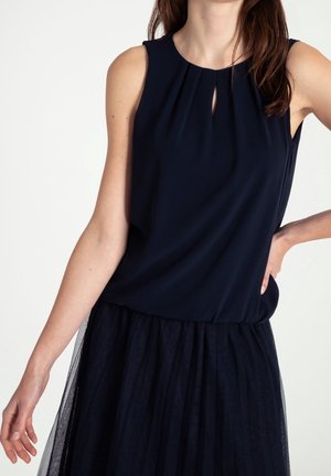 Woman wearing sleeveless black top with a small front keyhole and a layered black skirt with a tulle overlay against a white background.