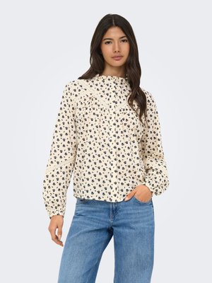 Femme aux longs cheveux foncés porte un chemisier crème avec un petit imprimé floral bleu marine et des détails en dentelle, associé à un jean bleu, debout avec la main dans la poche.