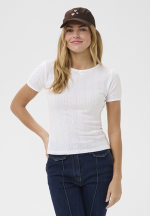 Young woman with long blonde hair wearing a brown cap with hearts, white short-sleeve patterned top, and dark blue jeans, posing with hand on hip.