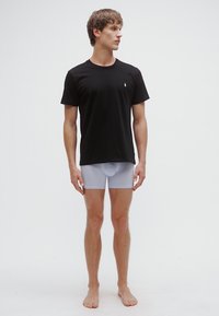 Camiseta preta de manga curta com um pequeno logótipo ao peito, combinada com cuecas boxer cinza claro, em pé descalço contra um fundo simples.