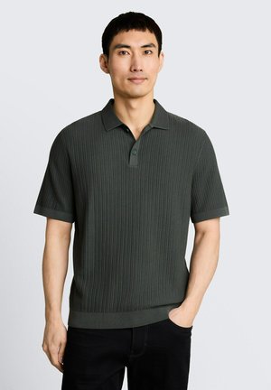 Homme aux cheveux courts noirs portant un polo à manches courtes en maille côtelée vert foncé et un pantalon noir, debout avec une main dans la poche.