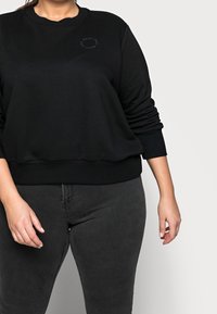 Zwarte sweatshirt met ronde hals, geribbelde mouwen en onderrand. Bevat een subtiele, verhoogde logo op de borst. Draagt met donkergrijze jeans.