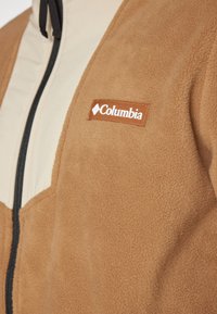 Brun fleecejakke med cremefarvede accenter, med lynlås foran og Columbia-logo i brun på brystet. Blød tekstur.