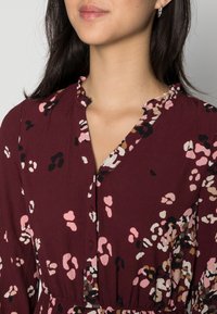 Burgundy klänning med en texturerad tyg, med V-ringning och knappar, prydd med ett blommönster i rosa, svart och kräm.