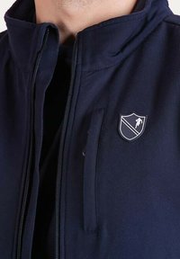 Veste zippée bleu marine avec un col haut, présentant un logo en forme de bouclier argenté et une poche latérale, fabriquée dans un tissu lisse avec une légère texture.