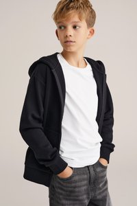 Fekete cipzáros kapucnis felső, puha textúrával, nagy kapucnival és elülső zsebekkel, egy fehér pólóra rétegezve, szürke farmernadrággal párosítva.