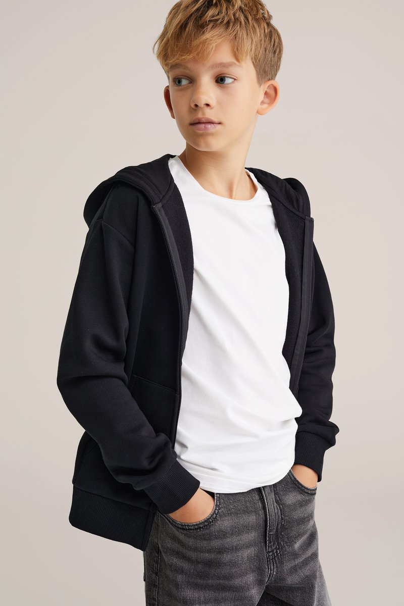 Fekete cipzáros kapucnis felső, puha textúrával, nagy kapucnival és elülső zsebekkel, egy fehér pólóra rétegezve, szürke farmernadrággal párosítva.