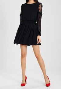 Černé šaty s dlouhými průhlednými krajkovými rukávy, volány na ramenou a přiléhavým pasem. V kombinaci s červenými vysokými podpatky.